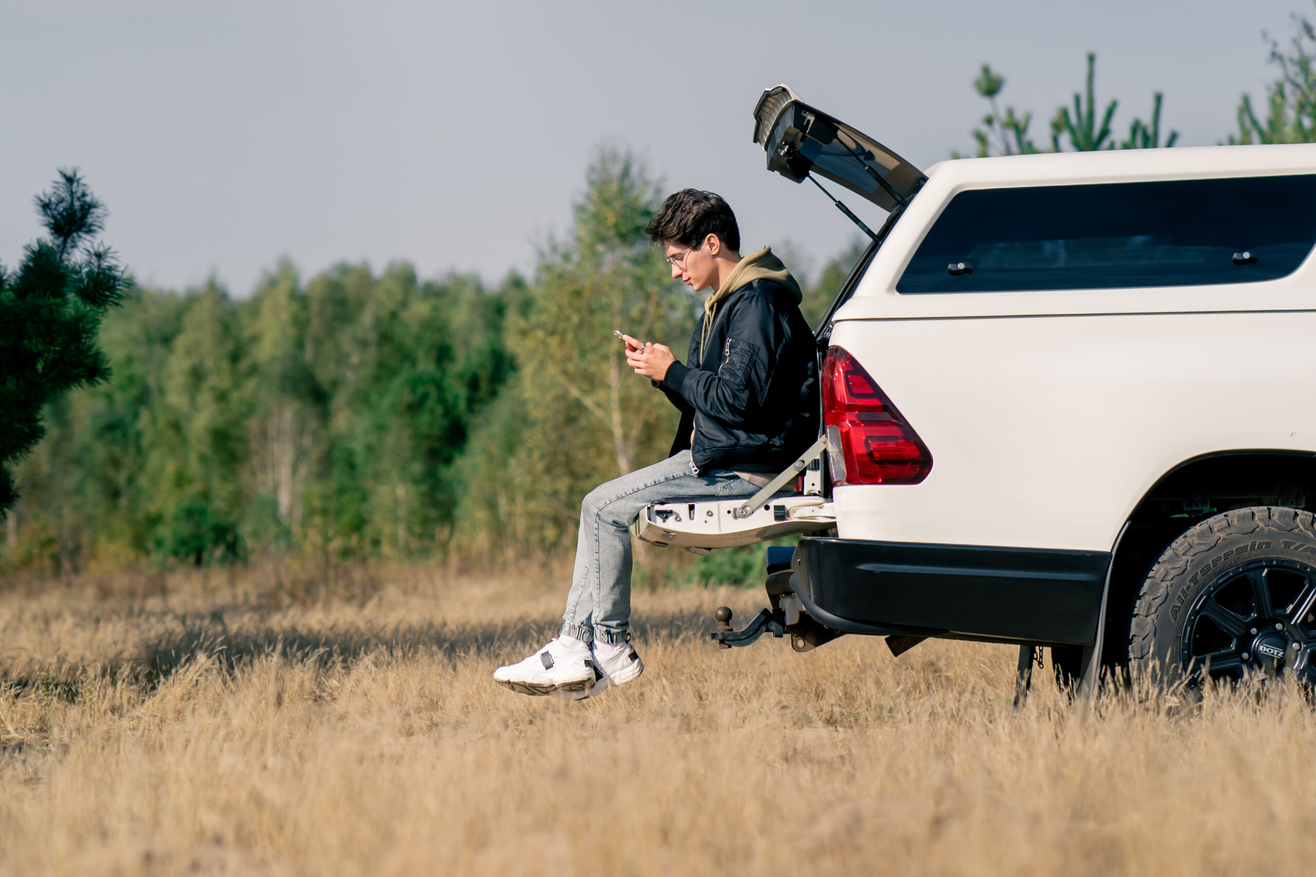A young guy sits in the open trunk of a car with phone and studies the area during a stop in meadow
